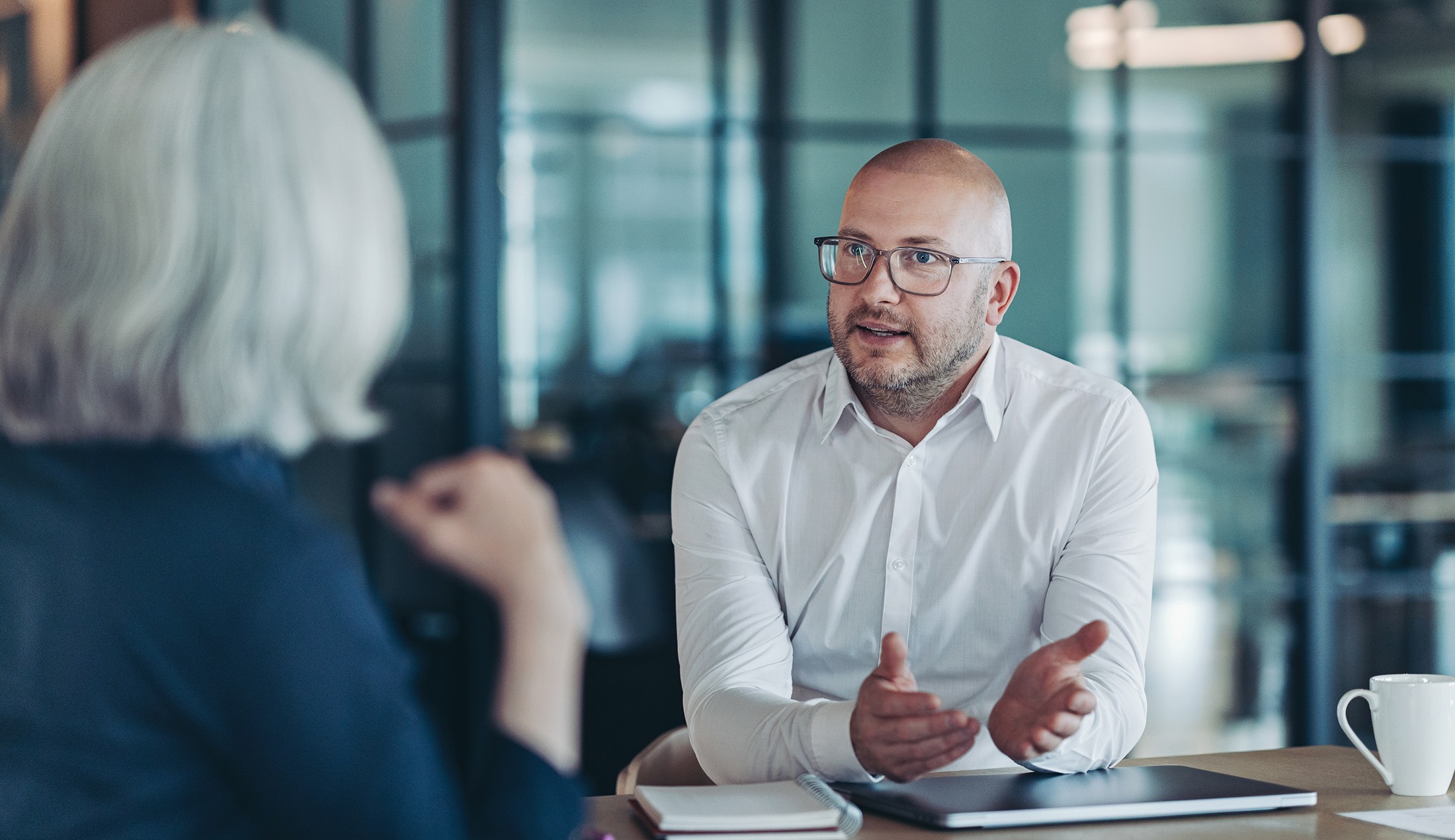 En man sitter vid ett skrivbord och pratar med en kvinna, vars rygg man ser.