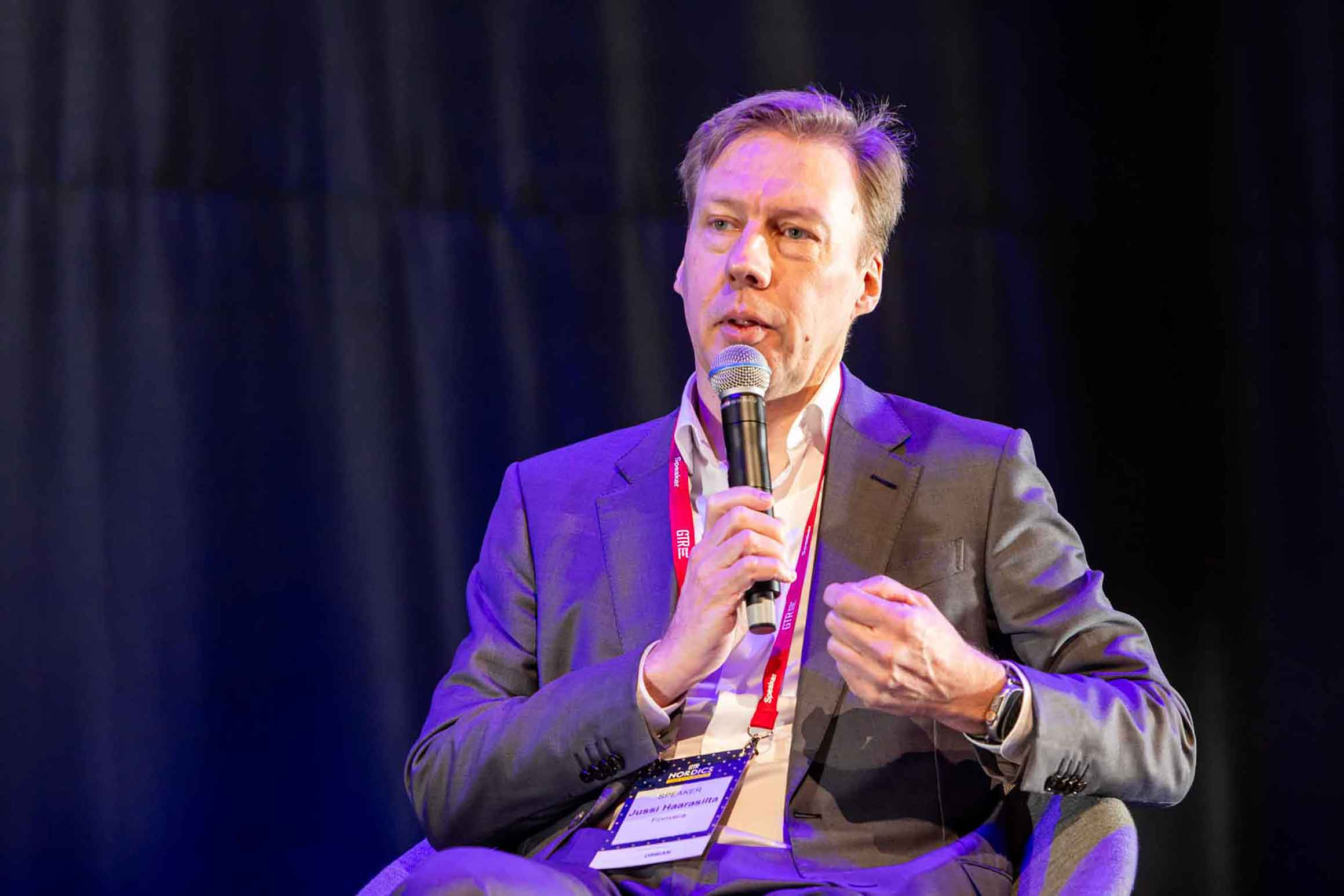 Jussi Haarasilta is sitting in an armchair on a stage, holding a microphone.