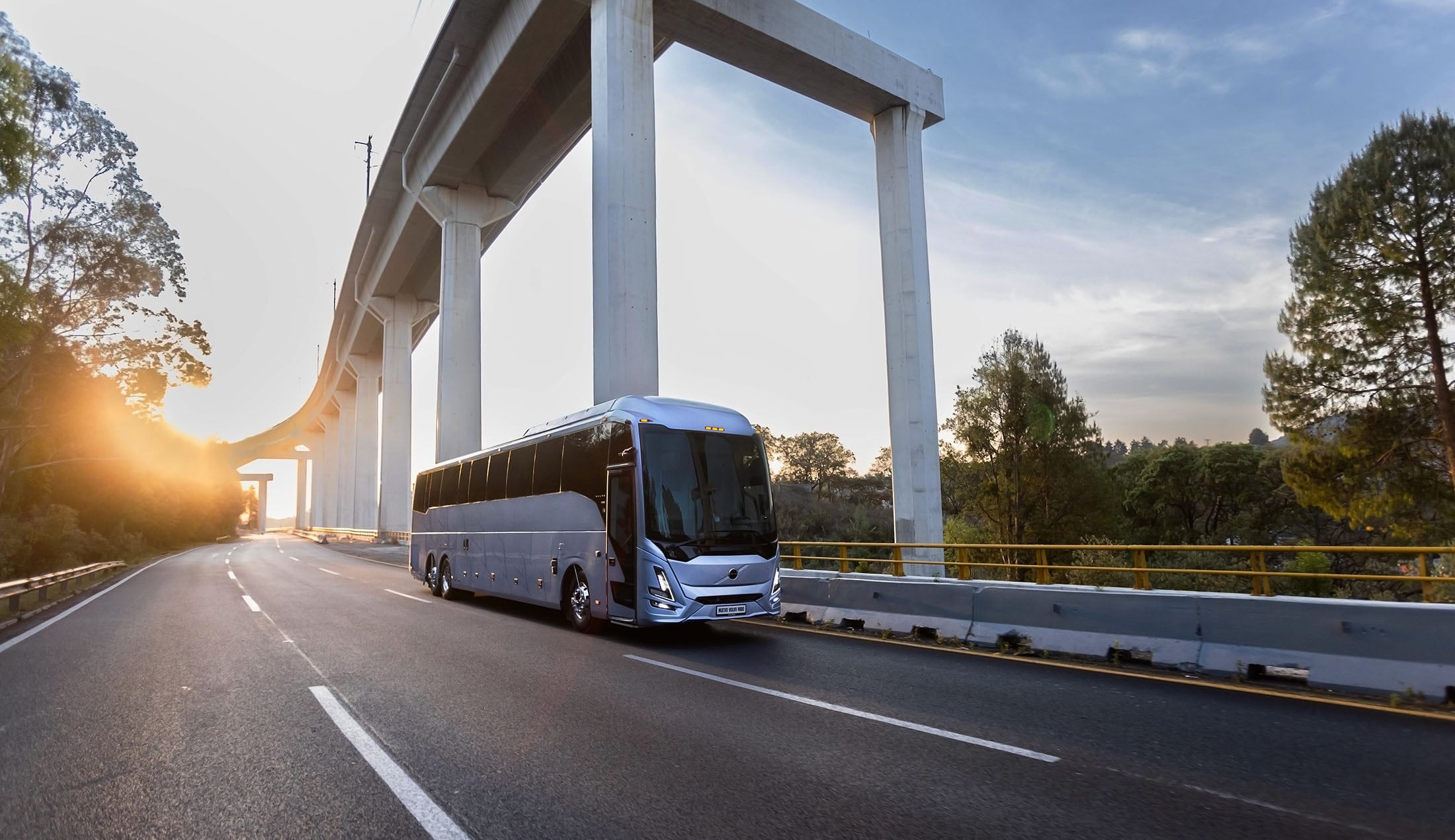 A silver coach bus travels along a highway in soft morning or evening light.
