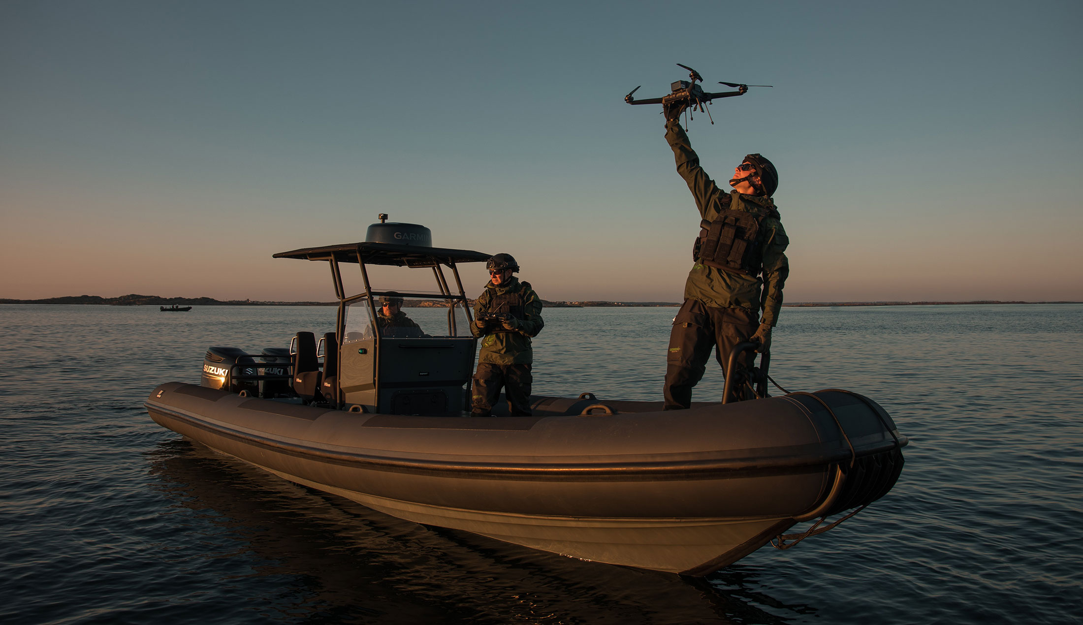 Tre militärer i en stor gummibåt. Mannen som står i fören håller upp en drönare mot skyn.