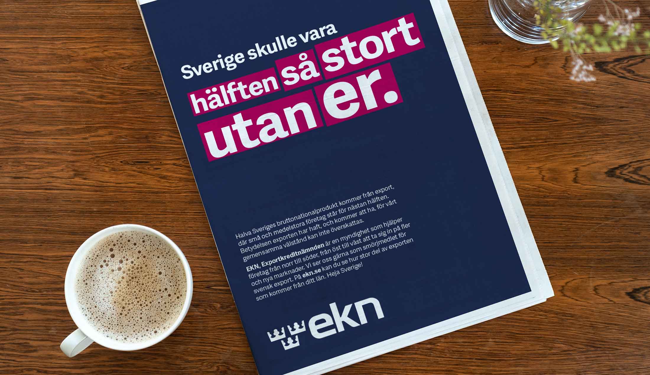 På ett bord ligger en tidning med en annons för EKN med texten "Sverige skulle vara hälften så stort utan er." På bordet står också en kaffekopp och en blomvas skymtas.