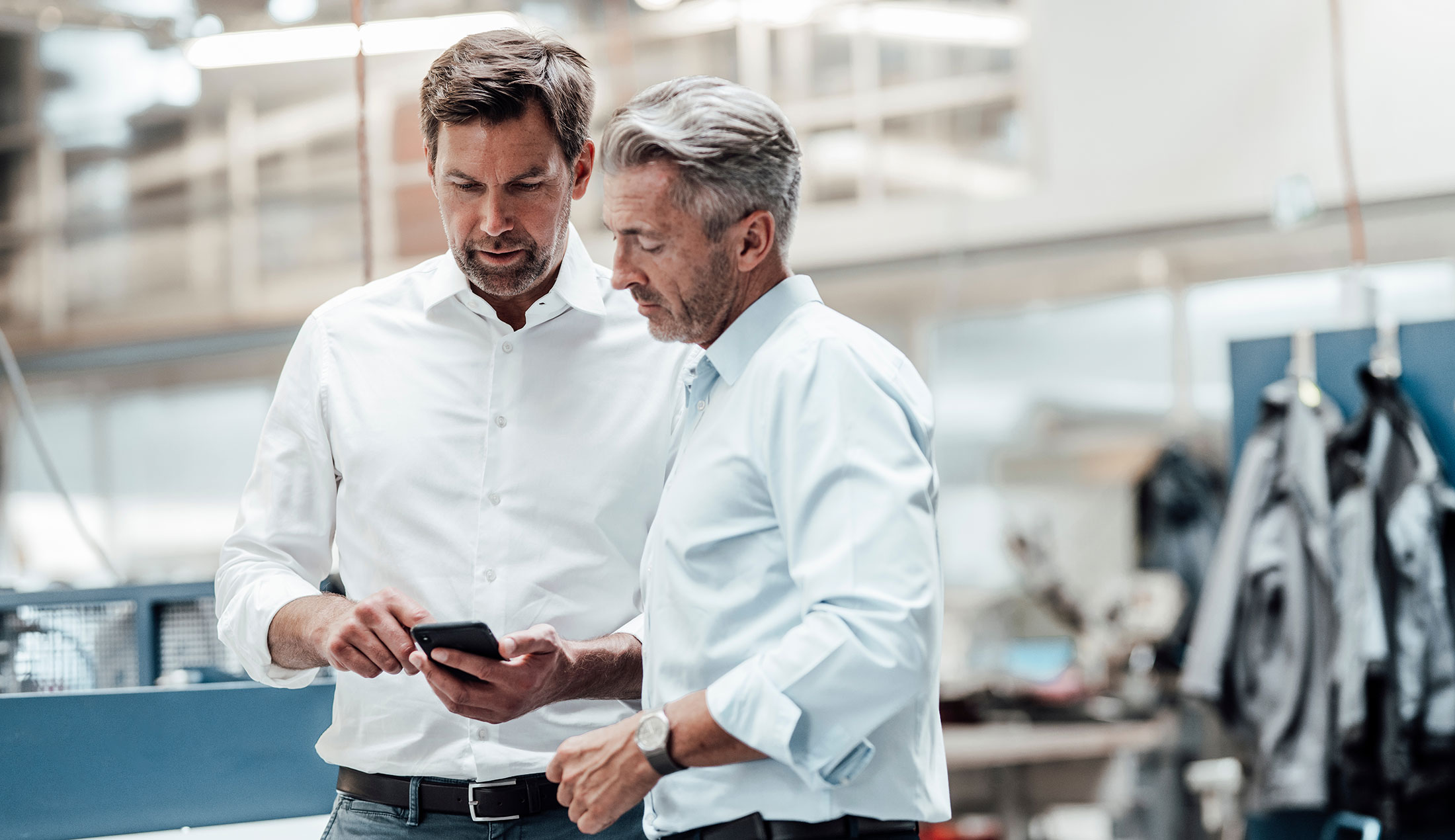 ChatGPT sade:  Two men standing in a bright industrial setting, looking at one of the men’s mobile phone.