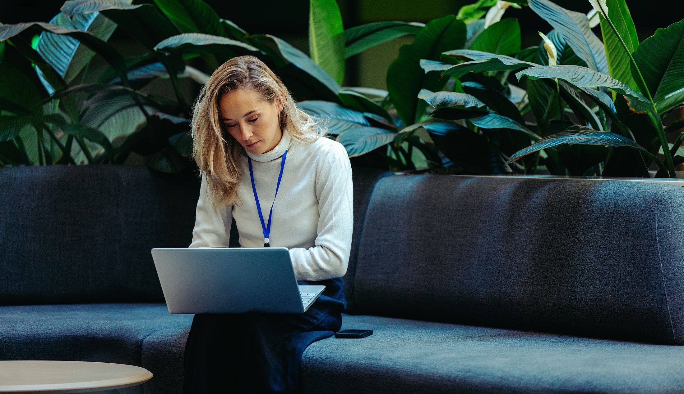 A woman sitting on a sofa with her laptop on her lap. Lush plants can be seen in the background.