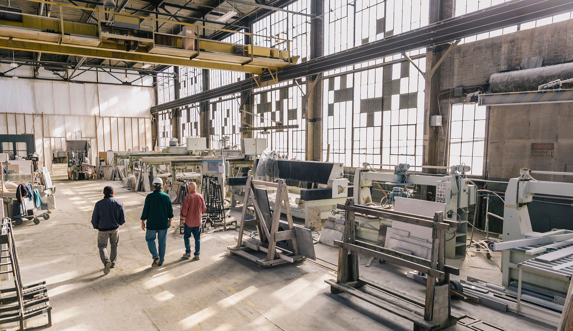 Three people walking through a bright factory building with large windows.