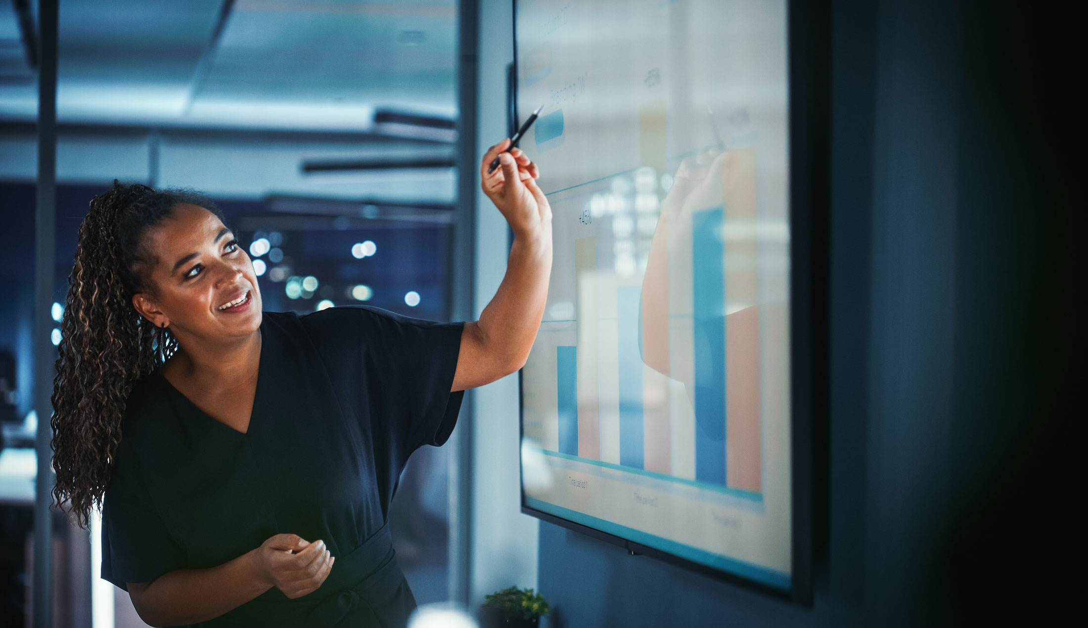 A woman standing in an office environment, pointing to something on a chart displayed on a wall-mounted screen.