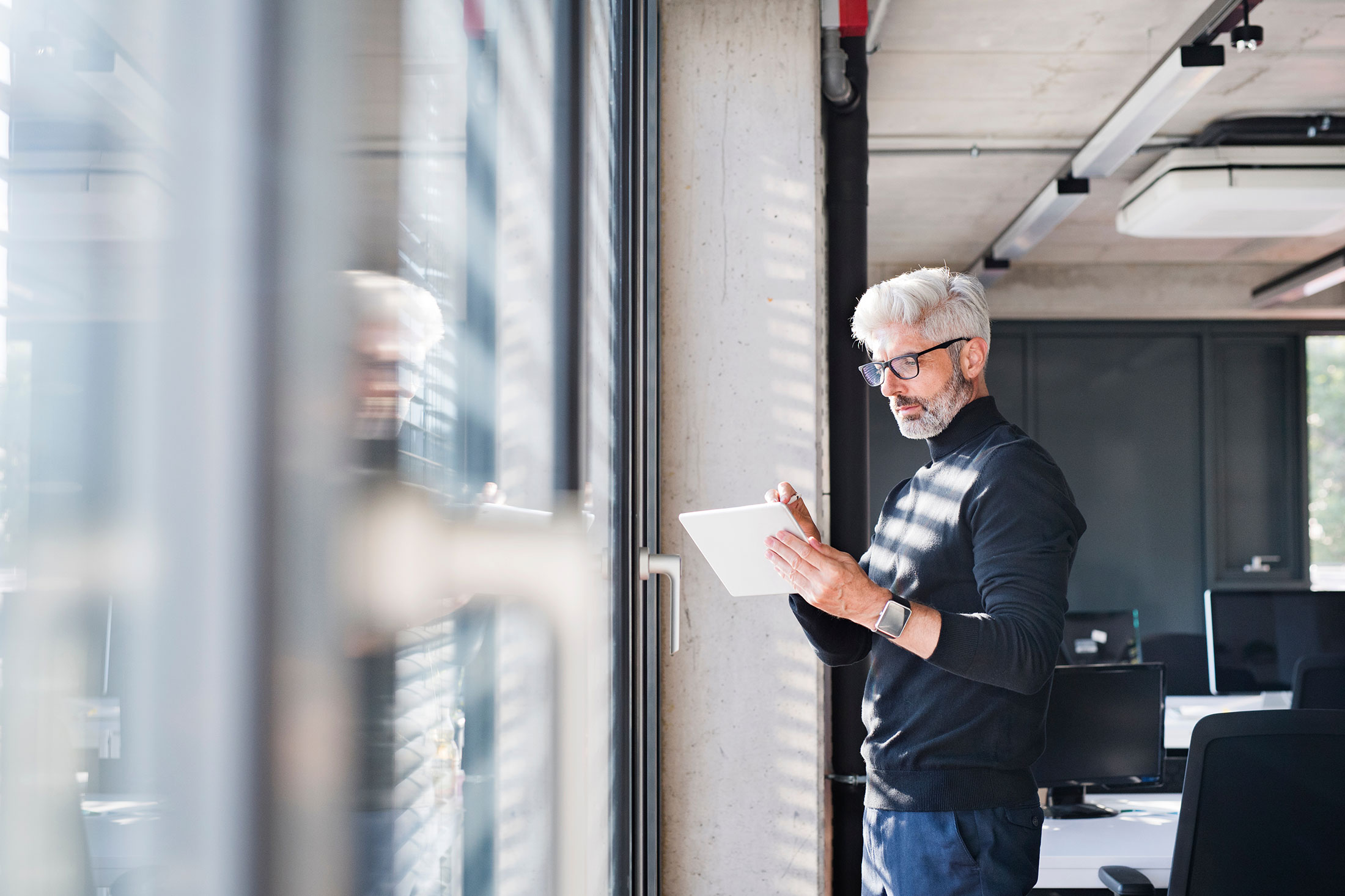 A man stands by an office window, reading on his tablet.